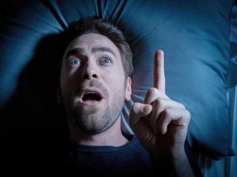 Shocked man lying in bed at night pointing finger up with sudden realization and surprise