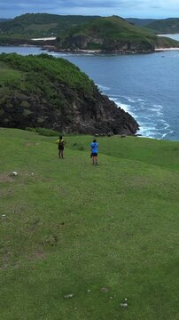 Couple Walking On Grassy Hill Overlooking Turquoise Ocean, Aerial Drone Shot Captures Sweeping Shoreline And Rolling Green Ridge. Two Travelers Stroll Along Winding Footpath Leaving Faint, Lombok