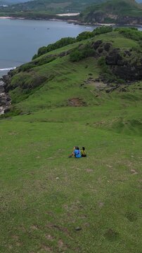 Couple sitting grassy hill overlooking ocean, asian pair wearing blue and yellow backpacks, watching rocky cove and calm sea, panoramic green slopes, intimate romantic retreat, Lombok, Vertical video