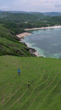 Couple Walking On Grassy Hill Overlooking Turquoise Ocean, Aerial Drone Shot Captures Sweeping Shoreline And Rolling Green Ridge. Two Travelers Stroll Along Winding Footpath Leaving Faint, Lombok