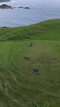 Couple sitting grassy hill overlooking ocean, asian pair wearing blue and yellow backpacks, watching rocky cove and calm sea, panoramic green slopes, intimate romantic retreat, Lombok, Vertical video