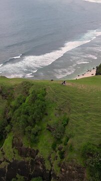 Group on cliff overlooking ocean, three hikers pause on grassy headland, aerial view reveals rolling waves, surf lines, distant islands and sandy bay under moody clouds,Bukit Merese, Lombok, Indonesia