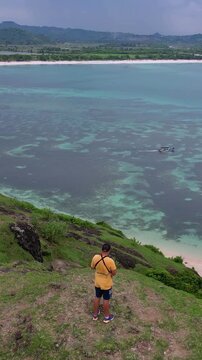 Man overlooking turquoise ocean from cliff, lone traveler and photographer stands on grassy headland, aerial drone perspective reveals coral reef patches, sandy bay and distant boats, Lombok