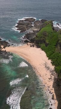 Aerial tropical beach cove with turquoise water and rocky headland waves breaking over coral reef, lush green vegetation cascading down cliffs, Bukit Merese, Lombok,Tanjung Ann, Indonesia