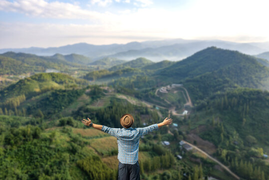Rear view of male traveler with raised arms standing on mountain peak enjoying beautiful nature landscape.