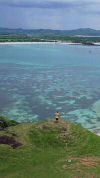 Man overlooking turquoise ocean from cliff, lone traveler and photographer stands on grassy headland, aerial drone perspective reveals coral reef patches, sandy bay and distant boats, Lombok
