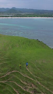 Couple Walking On Grassy Hill Overlooking Turquoise Ocean, Aerial Drone Shot Captures Sweeping Shoreline And Rolling Green Ridge. Two Travelers Stroll Along Winding Footpath Leaving Faint, Lombok