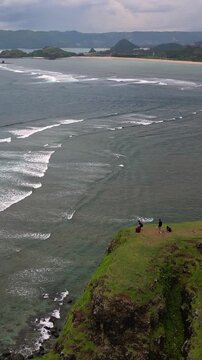 Group on cliff overlooking ocean, three hikers pause on grassy headland, aerial view reveals rolling waves, surf lines, distant islands and sandy bay under moody clouds,Bukit Merese, Lombok, Indonesia