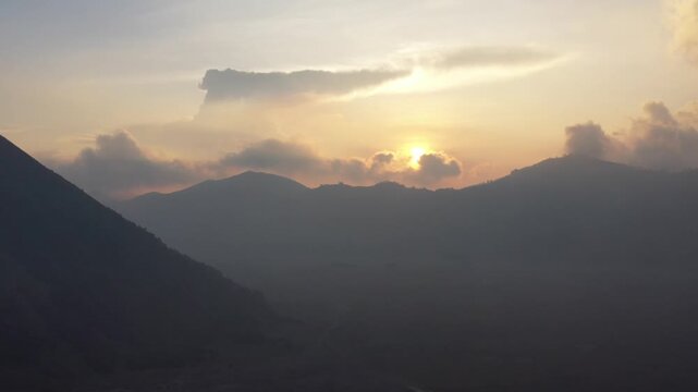 Drone pans left from an orange sunrise behind clouds to reveal silhouettes of Mount Batok and smoking Mount Bromo, East Java, Indonesia. Cinematic volcanic sunrise b-roll.