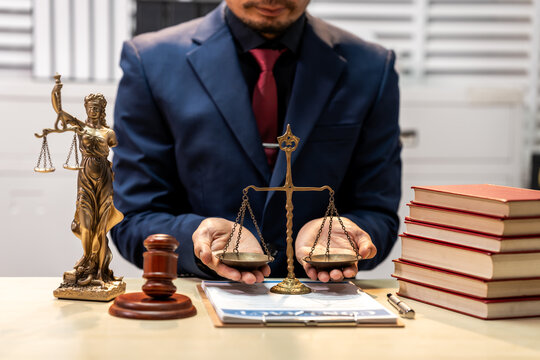 Lawyer in suit holding scales of justice with gavel, law books and documents on desk, legal office scene showing fairness and legal consultation