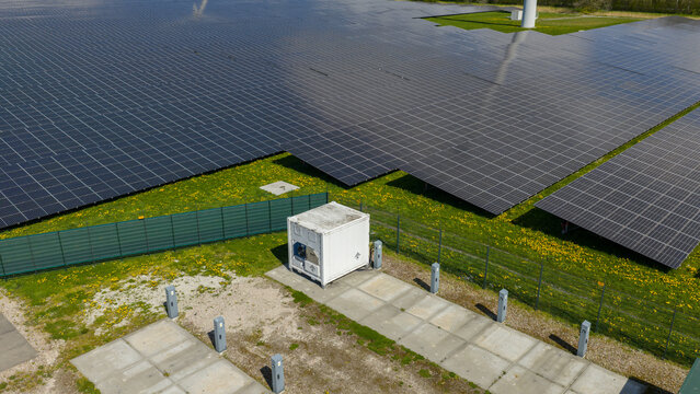 Aerial view of a large solar farm with rows of photovoltaic panels and a white electrical substation container in Opmeer, North Holland, Netherlands.