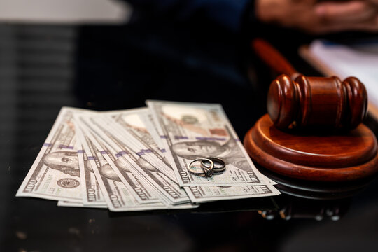 Wedding rings and us dollar bills beside a judge gavel, showing divorce, settlement and legal money issues.