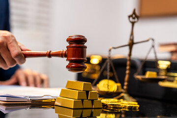 Judge gavel striking near gold bars, coins and scales in a legal finance concept with blurred courtroom background © Thitaporn