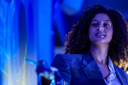 Businesswoman with curly hair outlines the roadmap with open hands rim lit in gold while speaking beside a deep blue wall inside a futuristic office illuminated by glowing blue projections