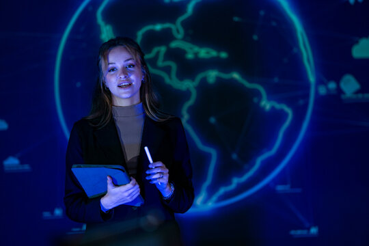 Young businesswoman with long fair hair pointing a stylus at a glowing world map wall highlights a region with the pen while the team records a technology explainer in the darkened studio