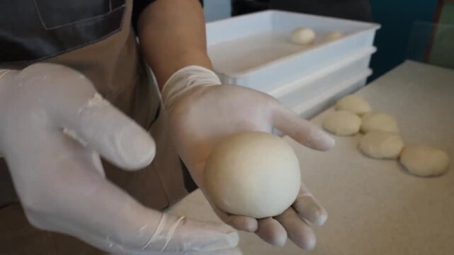 An experienced pizza maker (pizzaiolo) wearing white gloves expertly hand-rolls and shapes raw yeast dough into smooth balls on a floured countertop. This high-quality footage showcases the traditiona