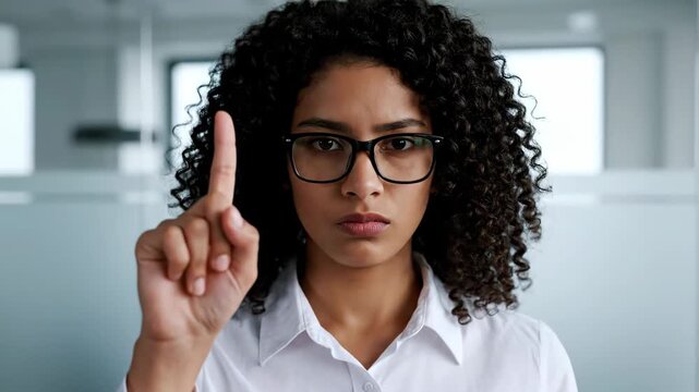 Serious young woman in glasses raising index finger in warning or prohibition gesture.