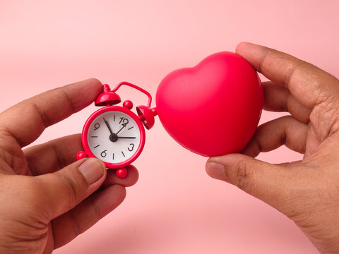 Two hands hold a small red mechanical alarm clock next to a matching red stress ball heart over pink