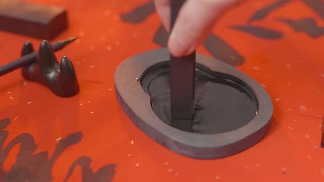 The calligrapher artist hands grinding stick ink on an inkstone ready to writes traditional chinese calligraphy on crimson paper, skillful brush strokes create elegant characters with wisdom writing