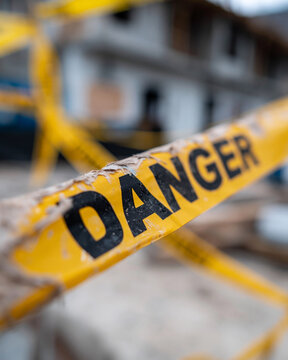 Yellow tape warns of hidden peril. Bold letters scream danger to all who approach. Construction site looms blurred in background. No people, just caution, urgency, and caution