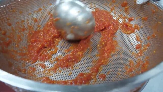 close-up shot of vibrant, crushed red tomatoes being strained through a metal sieve with a ladle. This process captures the traditional Italian method of preparing fresh pizza sauce or tomato pulp, en