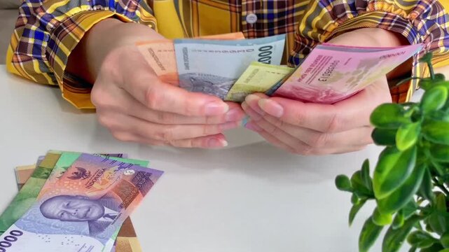 A close-up of a woman's hands counting Indonesian rupiah at her office desk. Suitable for topics such as finance, budgeting, money transfers, expenses, income, travel, and economics.