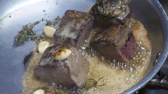 Close-up slow-motion footage of a professional chef using a spoon to baste thick beef tenderloin steaks with foaming melted butter, aromatic garlic cloves, and fresh rosemary in a stainless steel skil
