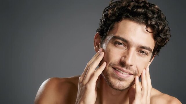 Close-up of a smiling curly haired adult man touching his cheeks with both hands against a plain gray background, lit with soft studio light and framed