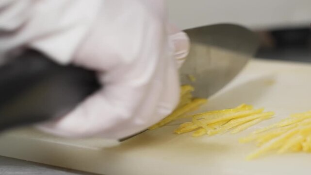 Close-up of a pastry chef's hands in white gloves using a sharp kitchen knife to precisely cut thin yellow lemon peel into julienne ribbons on a white cutting board. Ideal for culinary tutorials, fine