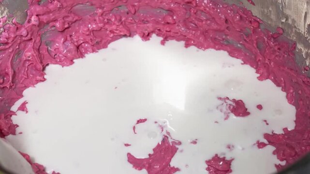 close-up, high-angle shot showing a stream of creamy white milk being poured into a bowl of thick, vibrant pink dessert batter. The batter, which has a textured and creamy consistency, appears to be f