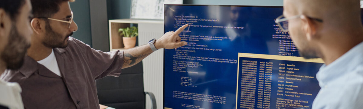 Young adult male developers reviewing source code on large monitor in information technology office, discussing software debugging and team workflow. For tech teamwork or coding process marketing