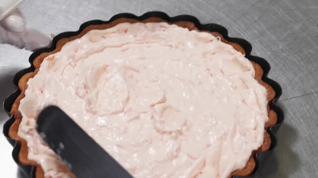 High-angle close-up footage of a pink strawberry or raspberry cream filling being smoothly spread into a golden-brown fluted tart shell using a white rubber spatula. The creamy texture of the dessert 