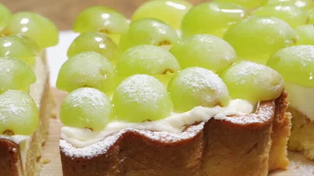 A close-up shot of a delicious homemade fruit tart featuring a golden-brown scalloped crust, smooth vanilla pastry cream filling, and a topping of juicy seedless green grapes dusted with powdered suga