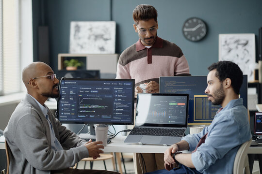 Multiethnic young adult men discussing software development workflow in information technology office, reviewing code and analytics dashboards. Ideal for teamwork, coding, startup marketing