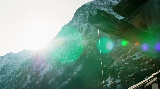Bright winter sun shines on a snowy mountain landscape, melting an icicle hanging from the wooden gutter of a rustic alpine cabin