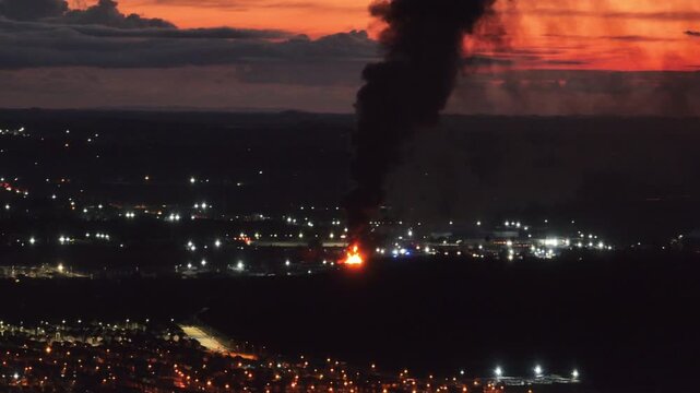 Drone zooms toward an industrial fire at night. Visible flames burn intensely as black smoke rises against a blood-red sky over a dark city below.