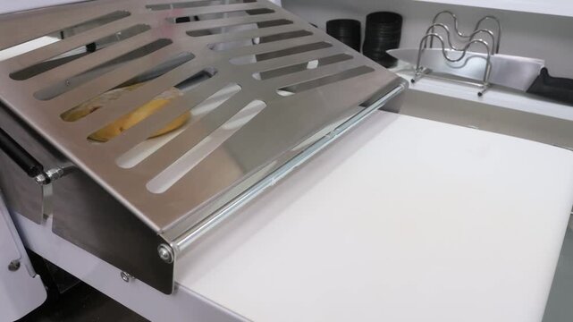 High-angle close-up of a commercial electric dough sheeter machine rolling out fresh yellow pastry dough on a conveyor belt. The stainless steel equipment accurately flattens the dough to a uniform th