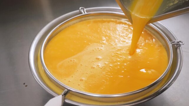 Close-up of vibrant orange-yellow custard or fruit curd being poured through a fine-mesh metal sieve into a bowl. This professional culinary process ensures a smooth, silky texture for artisanal pies,