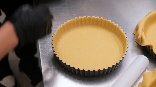 close-up, high-angle shot featuring a pastry chef wearing black gloves using a professional rolling pin to fit fresh dough into metal tart pans. The scene captures the artisanal process of preparing e