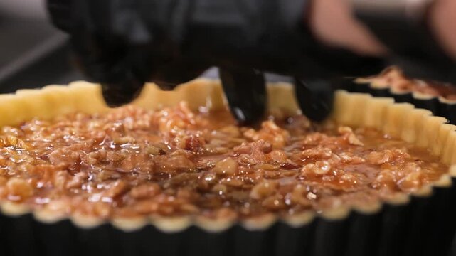Close-up of a pastry chef wearing a black nitrile glove carefully arranging walnut halves in a rich caramel-filled tart shell. The footage highlights the gooey texture of the caramel and the crunchy w