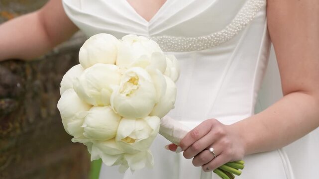 Elegant close-up shot of a bride in a white dress holding a lush peony bouquet outdoors, showcasing her engagement ring. Natural daylight and shallow depth of field enhance the romantic scene.