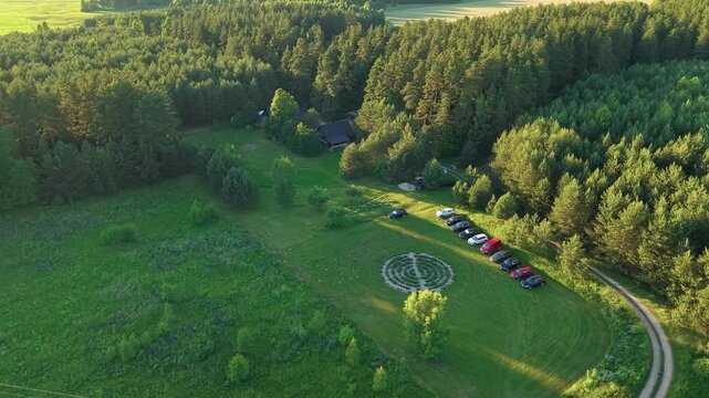 Drone video captures a wide aerial view of a rural clearing with parked cars, surrounded by dense green forest and sunlit meadows, on a bright, clear summer day.