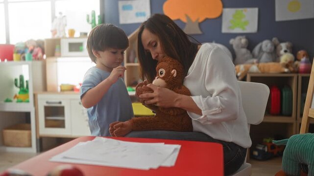 Woman seated holds a plush monkey while a little boy gently touches its paw and hands it a small toy over a low table in a building; bonding warmth nurturing.