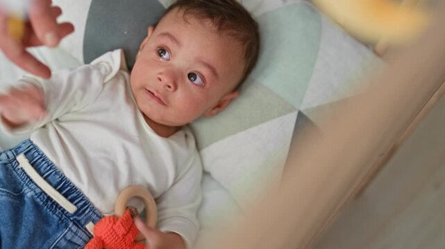 Baby boy crying in a crib, mouth open while hand offers a teether rattle near his face on a patterned mattress; distress need comfort.