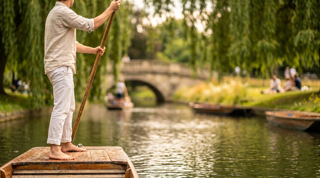 Barefoot man in white linen attire, holding long wooden punting pole, standing on flat bottom wooden boat, calm river canal with weeping willows and stone bridge, travel photography