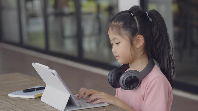 Young Asian girl with long hair and pink jacket using tablet device with headphones at table in modern indoor space, focused on screen