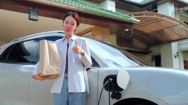 Asian female showing thumbs up sign and holding a paper bag near a charging electric vehicle at home. Concept of easy eco lifestyle and sustainable consumerism.