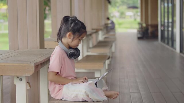 Child Girl Using Tablet Device Sitting on Floor in Modern Indoor Space with Large Windows and Wooden Interior, Casual Dress and Headphones, Relaxed Learning Environment