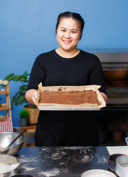 Asian happy chubby woman smile standing holding unbaked cacao liquid dough on tray being placed preparation stage process bakery showing handmade brownie cake before baking artisan bakery production.