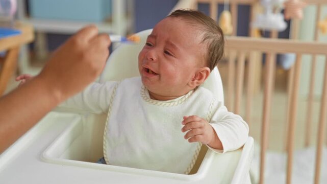 Hispanic baby boy crying with food on mouth and a spoon while seated in a nursery highchair, caregiver hand reaching to feed; feeding upset.